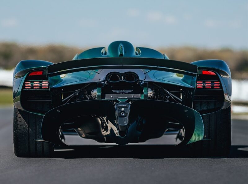 Rear view of a green Aston Martin Valkyrie Roadster on a road, showcasing wide tires, aerodynamic features, and exposed engine components in striking Green Carbon.