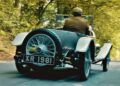 A person drives a vintage convertible car with the license plate XR 1981 along a tree-lined road, retracing the path of Valkyrie Meets Cloverleaf On Aston Hill: Where It All Began 100 Years Ago.