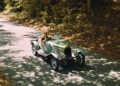 A person drives a vintage silver convertible car down a tree-lined road with dappled sunlight, evoking the spirit of Valkyrie Meets Cloverleaf On Aston Hill: Where It All Began 100 Years Ago.