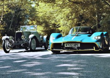 A vintage silver car and a modern blue sports car drive side by side on a tree-lined road in daylight—where Valkyrie Meets Cloverleaf On Aston Hill: Where It All Began 100 Years Ago.