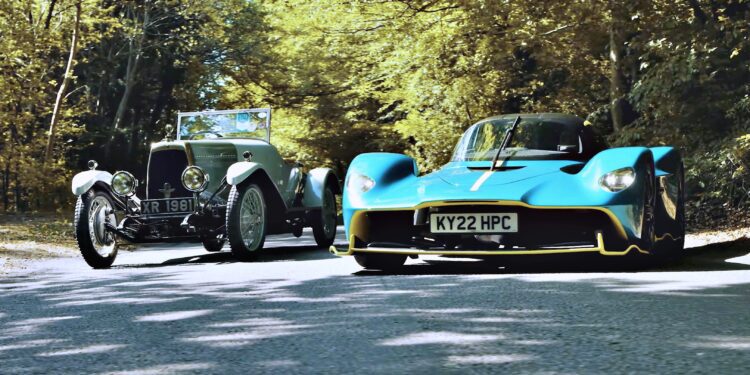 A vintage silver car and a modern blue sports car drive side by side on a tree-lined road in daylight—where Valkyrie Meets Cloverleaf On Aston Hill: Where It All Began 100 Years Ago.