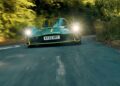 A green sports car with headlights on speeds around a tree-lined curve on Aston Hill, its "KY22 HPC" plate visible—where it all began 100 years ago: Valkyrie Meets Cloverleaf.