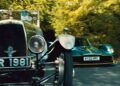 An old black car with license plate XR 1981 passes a modern green sports car, KY22 HPC, on a tree-lined road—Valkyrie Meets Cloverleaf On Aston Hill: Where It All Began 100 Years Ago.