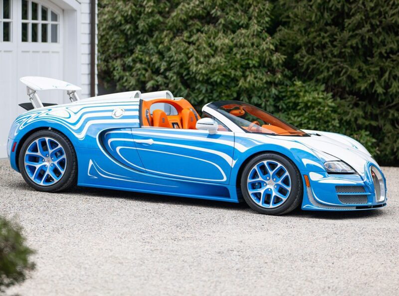 A rare 1-of-1 Bugatti Veyron Grand Sport Vitesse in blue and white with an orange interior sits on a gravel driveway, greenery and a building visible in the background.