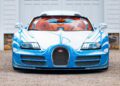 A front view of a blue and white Bugatti Veyron Grand Sport Vitesse with orange interior, parked on a gravel surface in front of a white building with large windows—a stunning 1-of-1 masterpiece.
