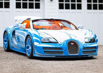 A blue and white Bugatti Veyron Grand Sport Vitesse convertible is parked on a gravel driveway in front of white garage doors.
