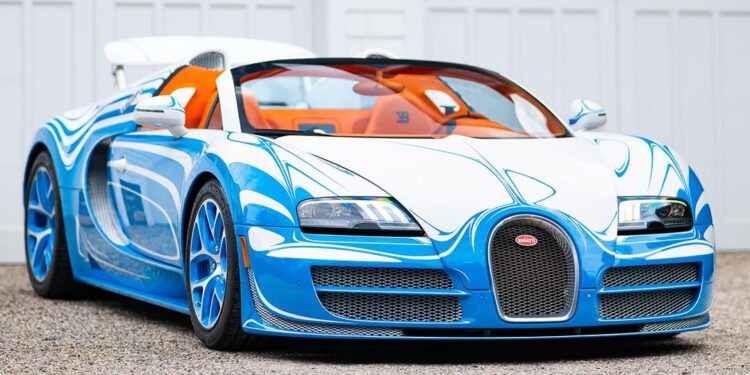 A blue and white Bugatti Veyron Grand Sport Vitesse convertible is parked on a gravel driveway in front of white garage doors.