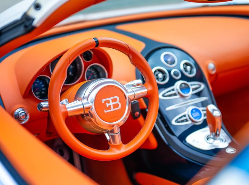 Close-up of a 1-of-1 Bugatti Veyron Grand Sport Vitesse interior featuring an orange steering wheel with the Bugatti logo, orange dashboard, and chrome-accented controls.