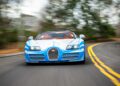 A blue and white Bugatti Veyron Grand Sport Vitesse, a rare 1-of-1 masterpiece, speeds down a curved road lined with blurred trees and houses.