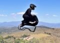 A person in a black suit and helmet is riding the Volonaut Airbike above a rural landscape with hills and mountains—no wings, no rotors: so how does this futuristic hoverbike fly?.