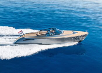 A modern motorboat with a tan and silver design moves quickly across calm blue water, offering a taste of the maritime experience that luxury yachts provide as it leaves a trail of white wake behind it.