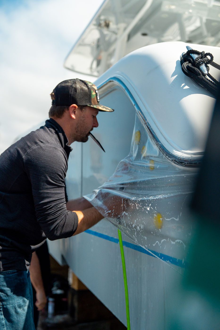 A man wearing a cap applies a clear protective film to the side of a boat while holding a tool in his mouth.