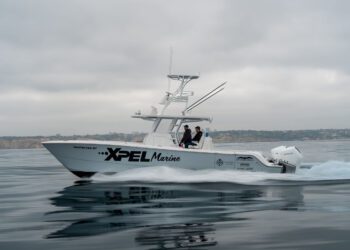 A white motorboat with "XPEL Marine" branding moves across calm water, with two people onboard and a coastline visible in the background under a cloudy sky.