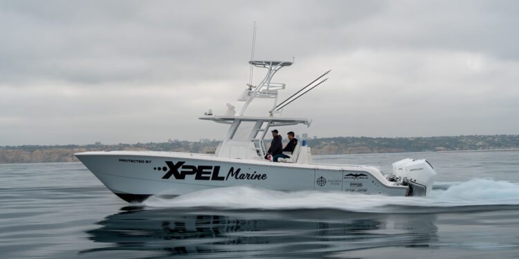 A white motorboat with "XPEL Marine" branding moves across calm water, with two people onboard and a coastline visible in the background under a cloudy sky.