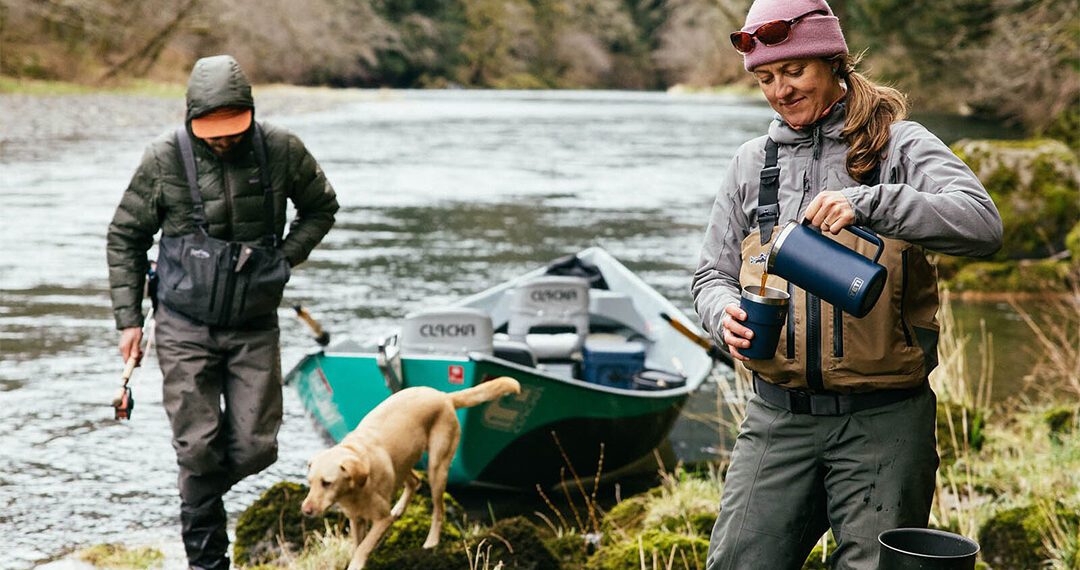 Two people and a dog are near a river with a drift boat; one pours a drink while the other holds a fishing rod. Surrounded by dense forest, they’re ready for an outdoor adventure with their essential gear.