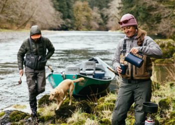 Two people and a dog are near a river with a drift boat; one pours a drink while the other holds a fishing rod. Surrounded by dense forest, they’re ready for an outdoor adventure with their essential gear.