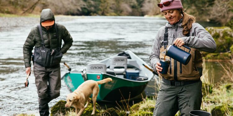 Two people and a dog are near a river with a drift boat; one pours a drink while the other holds a fishing rod. Surrounded by dense forest, they’re ready for an outdoor adventure with their essential gear.