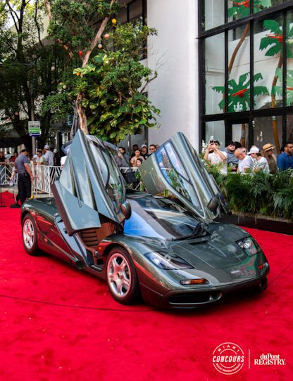 A metallic gray sports car with its butterfly doors open is displayed on a red carpet outdoors at the Miami Concours 2025, surrounded by a crowd and palm-themed windows—a true showcase of supercar rarity.