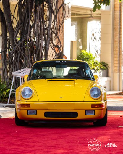 A yellow classic Porsche sports car, a true supercar rarity, is parked on a red carpet in front of a building with trees and outdoor furniture in the background.