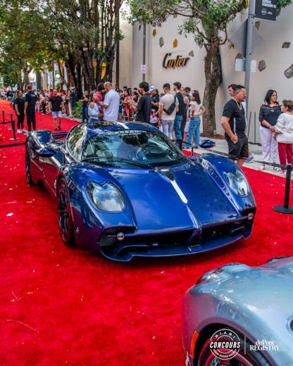 A blue sports car, showcasing supercar rarity, sits on a red carpet at an outdoor event near the Cartier store, with crowds and trees—capturing the luxury experience of Miami Concours 2025.