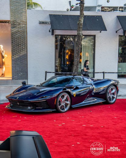 A black Ferrari supercar, symbolizing supercar rarity, is parked on a red carpet outside upscale shops, with a few people in the background.