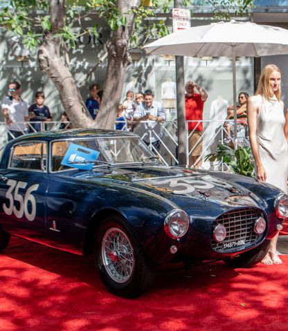 A classic dark blue car with the number 356 is displayed on a red carpet outdoors at the Miami Concours 2025. A woman in a white dress stands beside this supercar rarity, with onlookers in the background.