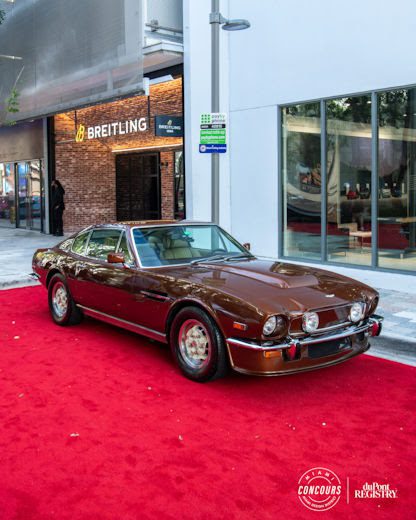 A vintage brown sports car is parked on a red carpet outside a Breitling store at Miami Concours 2025, with concours and duPont Registry logos highlighting the supercar rarity in this luxury experience.