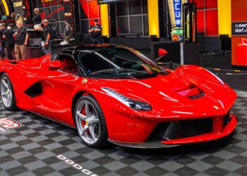 A red sports car is displayed indoors on a checkered floor, with several people and automotive displays in the background.