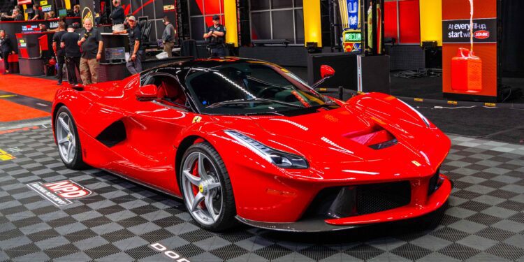 A red sports car is displayed indoors on a checkered floor, with several people and automotive displays in the background.