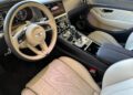 Luxury car interior featuring a beige leather seat, steering wheel with Bentley logo, digital displays, and a central control panel with various buttons and knobs.