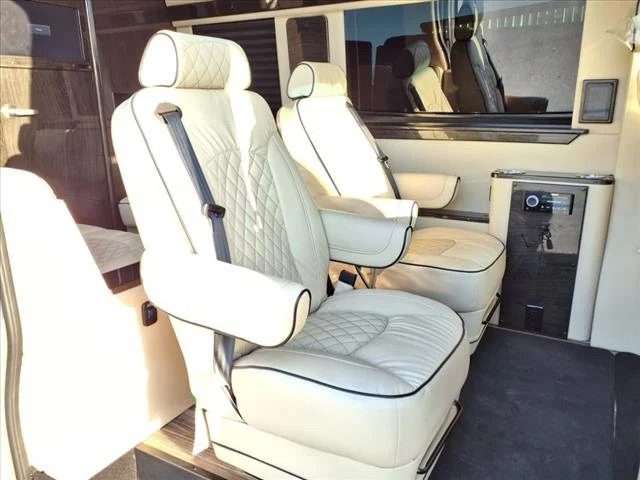 Two white leather seats with black trim inside a Custom Luxury Sprinter, featuring seatbelts, wood paneling, and a small safe mounted on the side wall—discover refined comfort in Ultimate Toys' Sprinters for sale.
