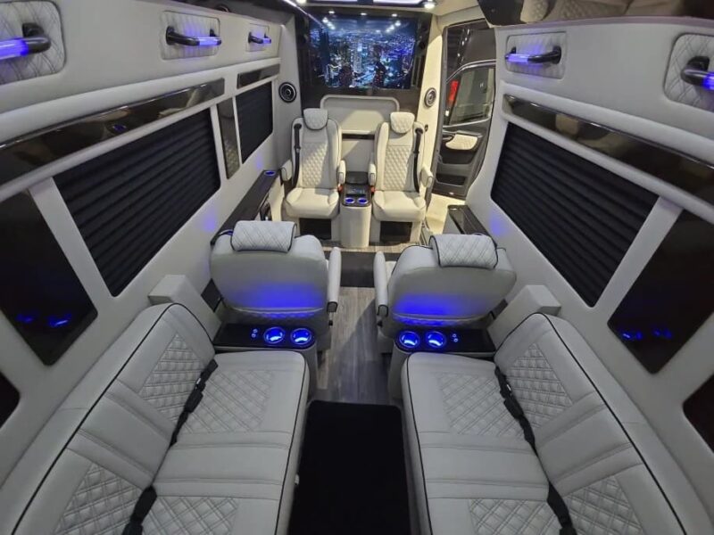 The image shows the interior of a custom luxury Sprinter with white quilted leather seats, blue accent lighting, cupholders, and a cityscape mural on the ceiling above the driver’s area—an Ultimate Toys masterpiece.