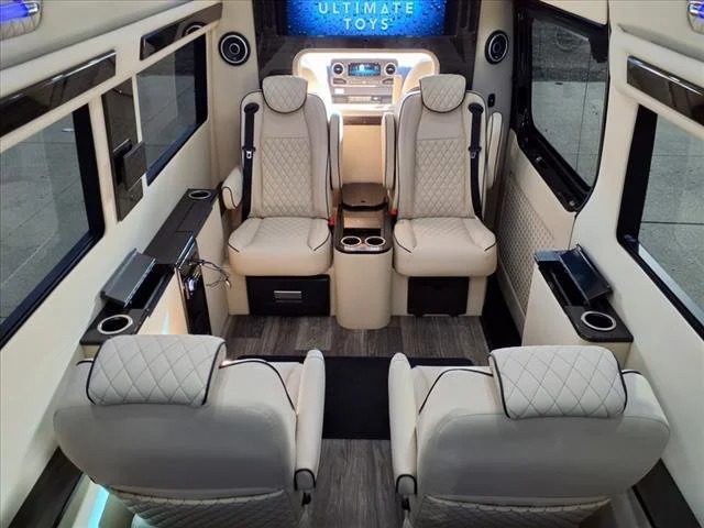 Interior of a custom luxury Sprinter van with four leather seats facing each other, cup holders, side tables, and a screen at the front labeled "Ultimate Toys." Discover more custom luxury Sprinters for sale from Ultimate Toys.