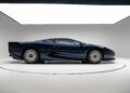 A Le Mans Blue Jaguar XJ220 supercar is displayed in a well-lit, white studio, shown from the side.