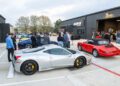 People gather outside a modern building with several sports cars, including a silver Ferrari and a red convertible, parked in the foreground on a concrete lot at an exclusive RAFA Racing Club event.