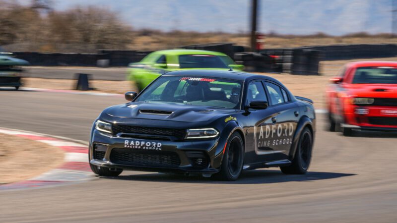 A black Radford Racing School car leads on a racetrack, followed by green and red cars, with a blurred background and dry landscape.