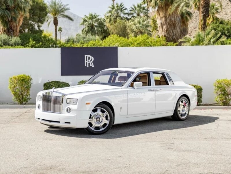A white Rolls-Royce Phantom sedan is parked on a paved surface in front of a white wall with the Rolls-Royce logo, surrounded by palm trees and greenery—an icon often favored by celebrity owners.