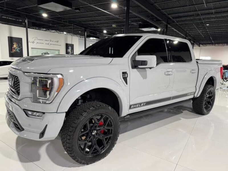 A white Ford F-150 Shelby pickup truck with black wheels and tinted windows is parked indoors on a glossy white floor, showcasing the bold design of Shelby Trucks.