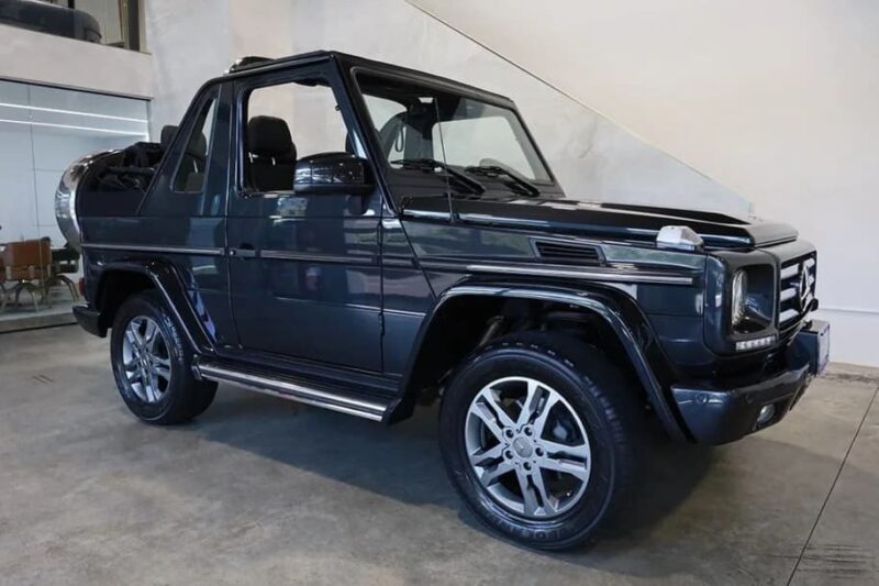 A dark gray Mercedes-Benz G-Class convertible luxury car is parked indoors on a concrete floor near white walls and glass partitions.