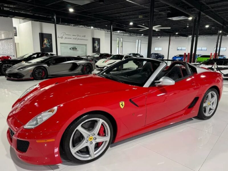 A red Ferrari convertible, an exotic car for sale, is parked indoors among several other luxury sports cars in a well-lit showroom.