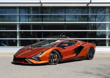 A metallic orange luxury car is parked outside a building with large glass windows, reflecting nearby trees.
