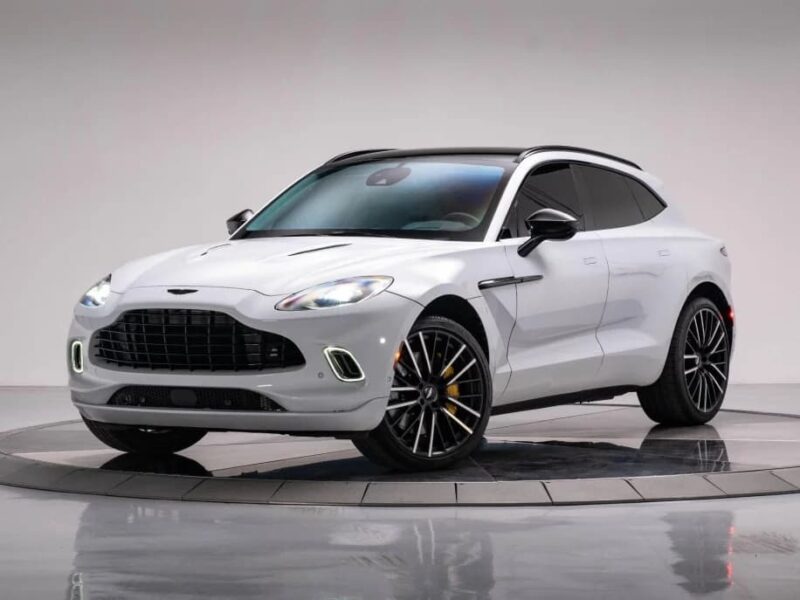 A white Aston Martin DBX luxury car is parked on a rotating display platform in a studio with a plain background.