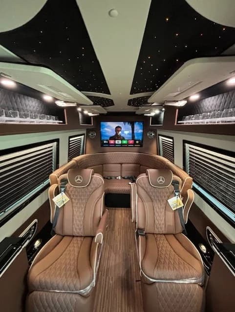 Luxury van interior with brown leather seats, wood flooring, LED star ceiling, and a large screen TV at the back. The Mercedes-Benz logo highlights this exotic car’s elegance.