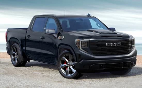 A black GMC pickup truck, a standout among trucks for sale, is parked on a gravel surface near a beach, with the ocean and cloudy sky in the background.