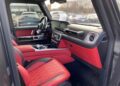 Interior of a luxury Mercedes-AMG G 63 featuring red and black leather seats, a digital dashboard, and modern controls, with cars visible through the window in a parking lot. This stunning BRABUS is now For Sale.