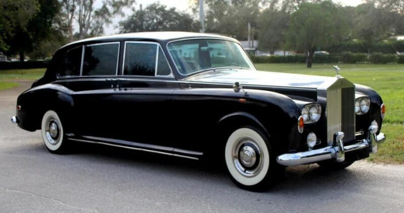 A classic black Rolls-Royce Phantom limousine with white wall tires, favored by celebrity owners for over 100 years, is parked on a paved road with grass and trees in the background.