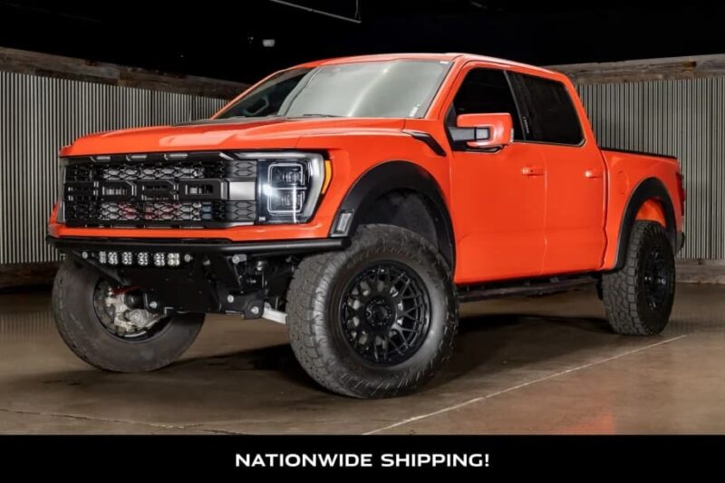 Bright orange lifted pickup truck with black accents and large off-road tires parked indoors on a concrete floor. Perfect rugged car for sale! Text at the bottom reads, "NATIONWIDE SHIPPING!.
