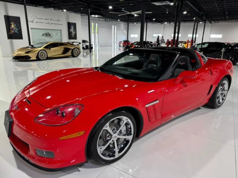 A red Chevrolet Corvette sports car is displayed in a showroom with other cars for sale, including a gold Lamborghini and other luxury car models in the background.