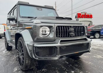 A gray BRABUS-modified Mercedes-AMG G 63 SUV is parked on a snowy lot near a dealership with a red Reina sign in the background, ready for sale.
