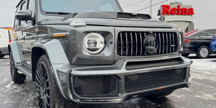 A gray BRABUS-modified Mercedes-AMG G 63 SUV is parked on a snowy lot near a dealership with a red Reina sign in the background, ready for sale.